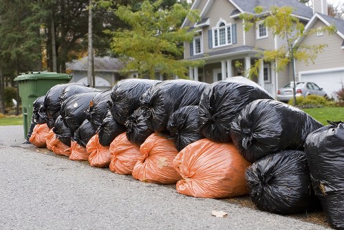 A row of tightly packed black and orange rubbish bags lined up along the edge of a residential street, with some bags leaning against a green wheeled bin. The black bags appear to contain general waste and are stacked neatly in front of the orange bags, which may contain lighter or different materials. The bags sit on an asphalt surface adjacent to a well-maintained driveway in front of a suburban property with a white house featuring large windows, a gable roof, and decorative brickwork. In the background, other houses, parked cars, and trees with yellow and green leaves are visible, suggesting a peaceful neighbourhood setting. The scene is illuminated by natural daylight, highlighting the textures of the plastic bags and the surrounding environment. This arrangement of waste items reflects typical rubbish collection preparation in the area and relates to professional waste removal services such as those provided by Commercial Waste Rickmansworth, specialising in waste management for local homes and businesses.