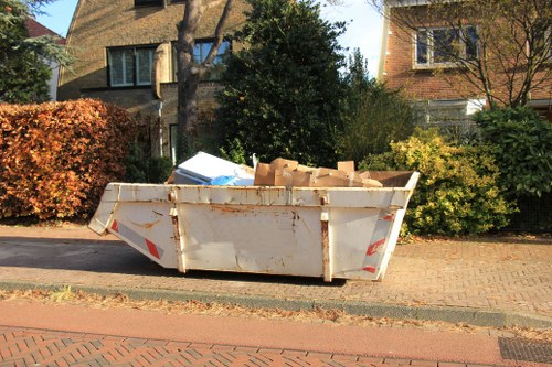 A large, weathered white metal skip filled with broken cardboard boxes and miscellaneous waste materials, positioned on a paved driveway adjacent to a residential street. The skip’s surface shows signs of rust and dirt, with some visible orange-brown corrosion near the base edges. The waste inside contains flattened cardboard boxes and packaging, with some edges protruding over the top. Behind the skip, there are green leafy bushes and trees, partially obscuring two brick and stone residential buildings with large windows and sloped roofs. The environment appears to be a quiet suburban area during daylight hours, with a clear sky casting natural light that highlights the textures of the waste and surroundings. This scene is representative of rubbish removal services available in the Rickmansworth area, facilitated by Commercial Waste Rickmansworth, supporting local waste management needs.