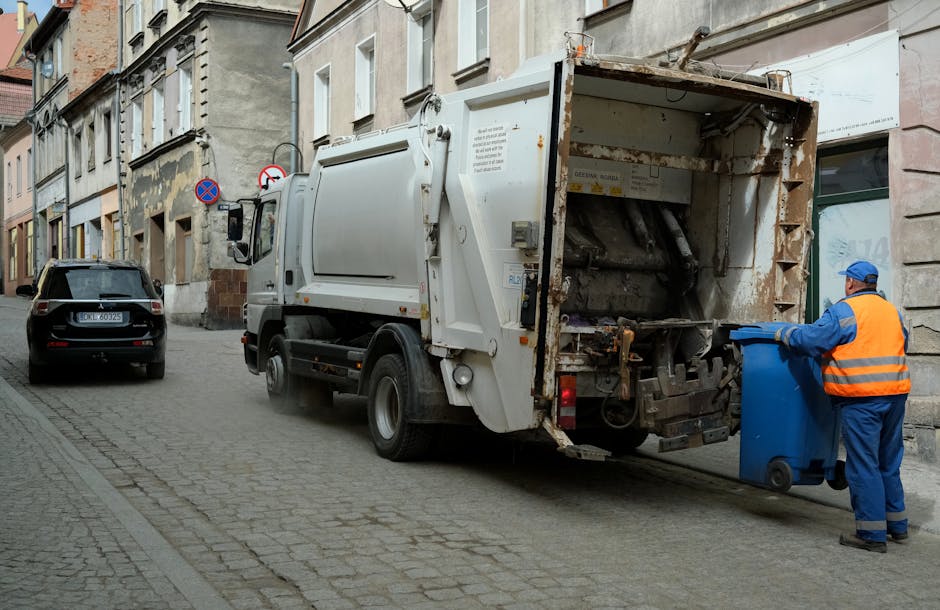 A person's hand is placing a crumpled brown paper bag into an open grey rubbish bin, which is partially visible in the background. The bin has a black interior and is situated on a wooden surface, wit